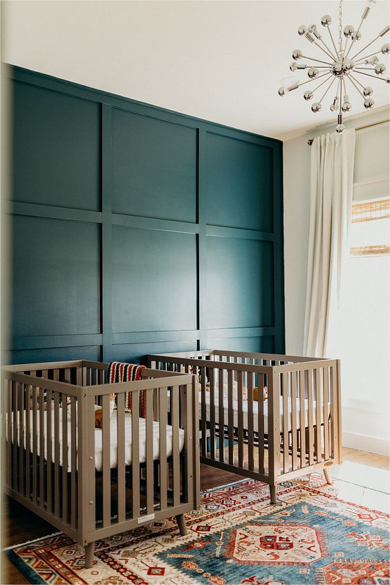 a bold twin nursery with a navy paneled wall, neutral cribs, a disco ball chandelier and a bright printed rug