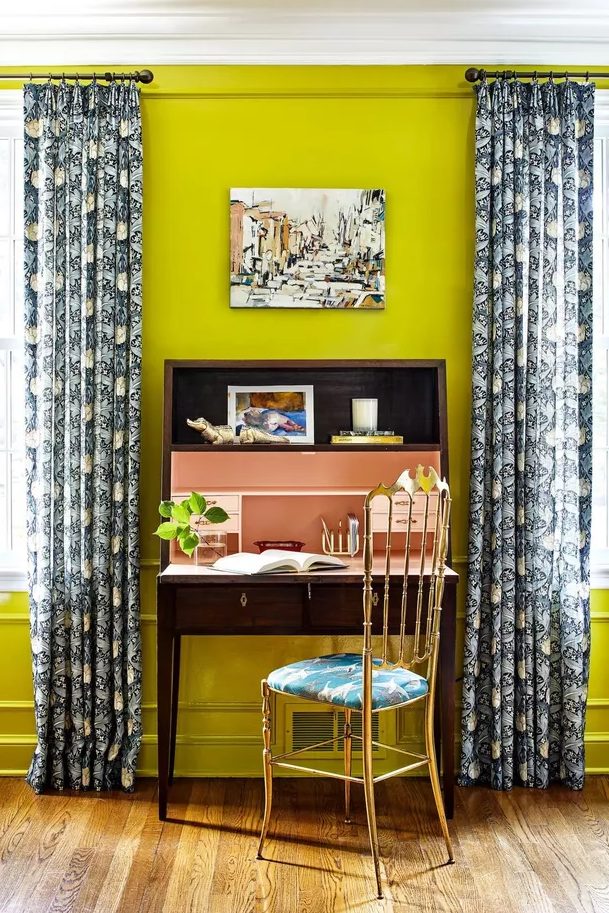 a bright home office with neon yellow walls, a vintage dark stained desk, a gilded chair with a tropical seat and printed curtains