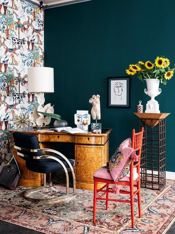 a bright maximalist home office wiht a teal and a wallpaper wall, a vintage desk, a navy chair and a red one, colorful textiles and a creative lamp
