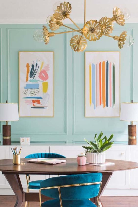 a bright maximalist home office with aqua panels, an oval desk and teal chairs, a gold floral chandelier and pretty artworks
