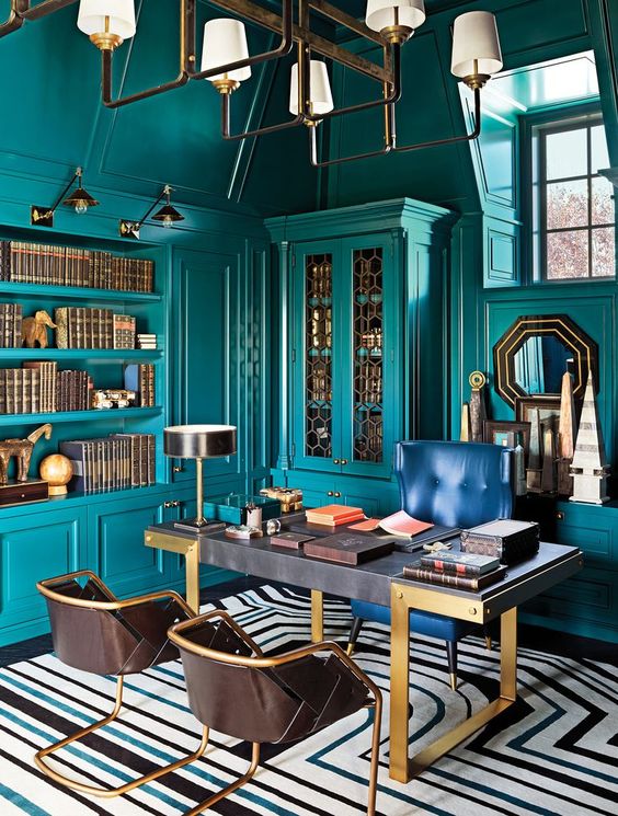 A bright maximalist home office with teal walls and matching built in furniture and shelves, a refined desk, a navy and leather chairs, a chic chandelier