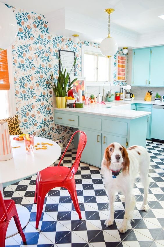 a bright maximalist kitchen with botanical wallpaper, light blue cabinets, a white table and red chairs plus a checked floor