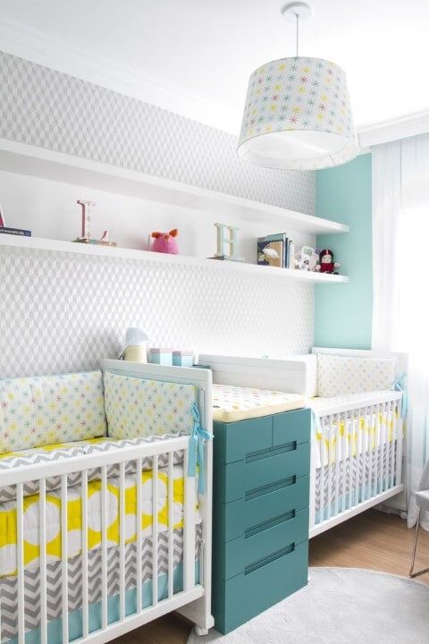 a bright twin nursery with an accent wall, white cribs, a teal changing table with storage, a pendant lamp and a shelf for display