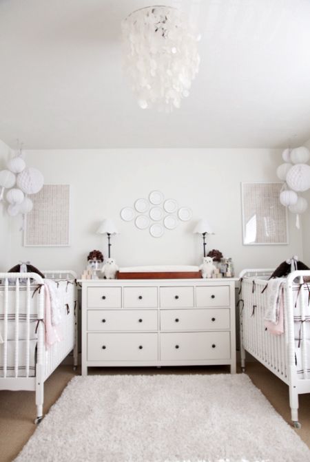 a chic twin nursery in white, with white furniture, neutral artworks, a mother of pearl chandelier and lovely bedding