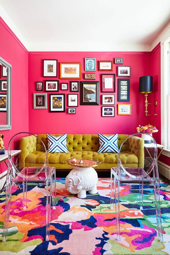 a colorful living room with hot pink walls, a mustard sofa, a colorful rug, acrylic chairs and an elegant gallery wall