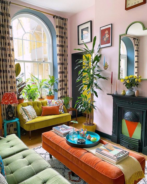 a colorful maximalist living room with bright furniture, a non-working fireplace, statement plants, printed curtains and pillows