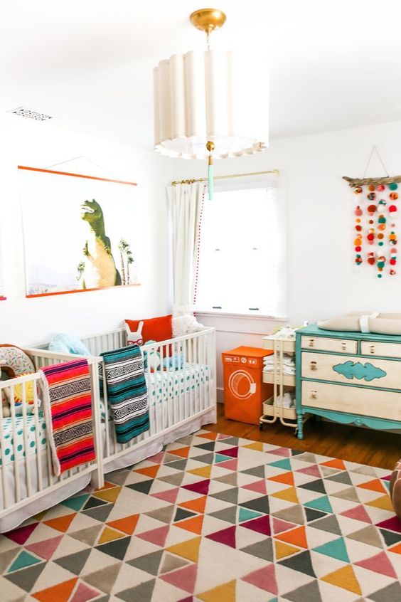 A colorful mid century modern nursery with white cribs, a neutral and turquoise dresser, colorful bedding and a colorful rug plus a cool chandelier