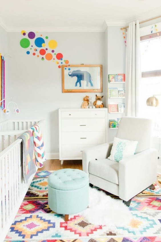 a colorful twin nursery with two white cribs, a dresser, a white chair and a turquoise ottoman, colorful textiles and decor