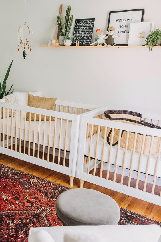 a cool boho twin nursery with white cribs, a Persian rug, a grey chair with a footrest, a shelf with some pretty pieces