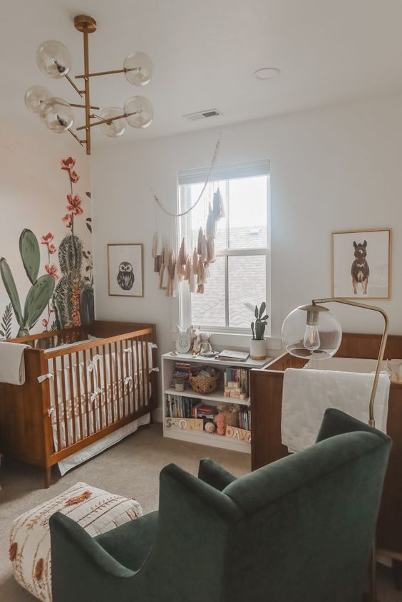 a desert themed twin nursery with stained cribs, cactus on the wall and dresser, a catchy chandelier and a green chair