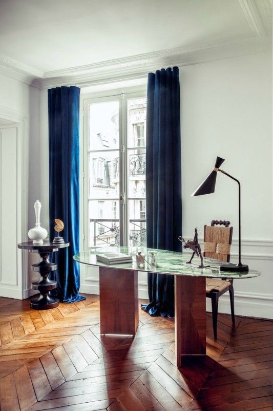 a fancy home office with a beautiful wood and marble desk, a woven chair, a black side table and navy curtains
