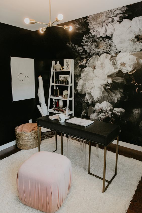 a fancy home office with a dark floral wall, a black and brass desk, a clear chair, a pink pouf and a basket plus a ladder shelving unit