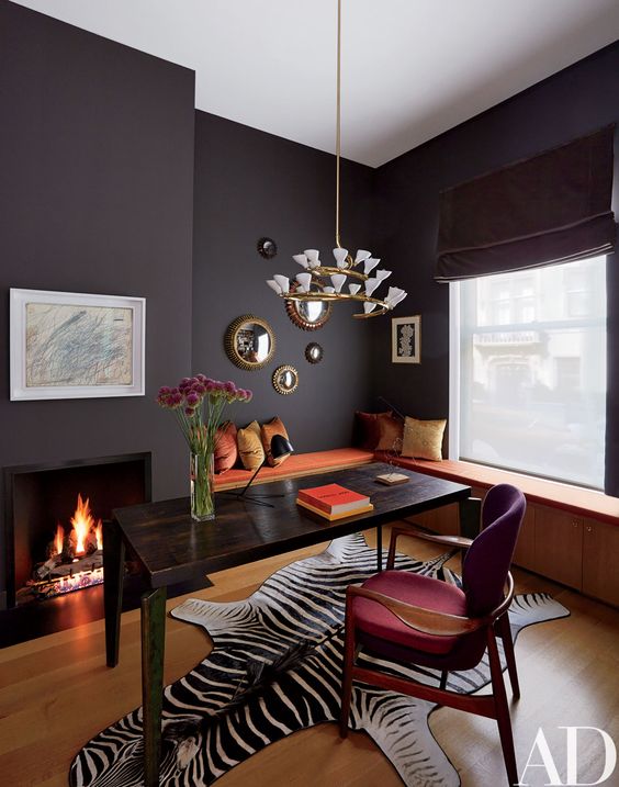 A fancy home office with a storage windowsill seating, a black desk, a fuchsia chair, a built in fireplace and a lovely chandelier