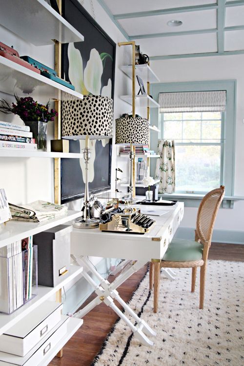 a fancy home office with light blue and white walls, a refined trestle desk, a rattan chair, open storage units and a statement floral artwork
