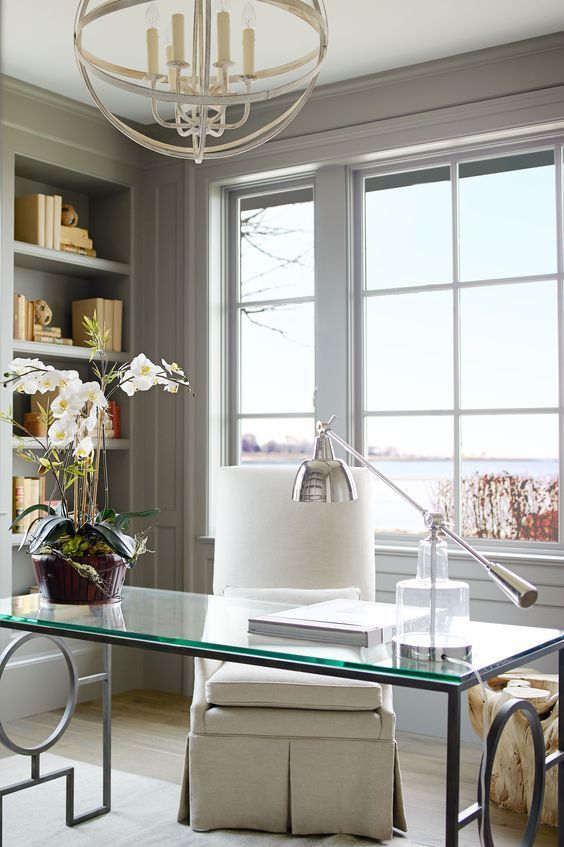 A fancy home office with light grey paneling, a glass desk and a creamy chair, built in shelves and a sphere chandelier