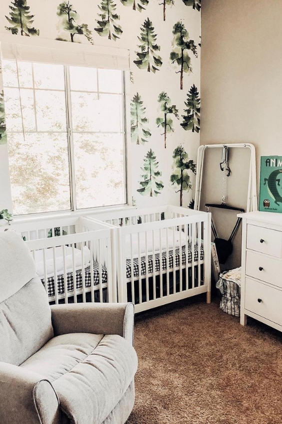 A forest themed twin nursery with white cribs, tree printed wallpaper, white and grey furniture and a pretty soft rug