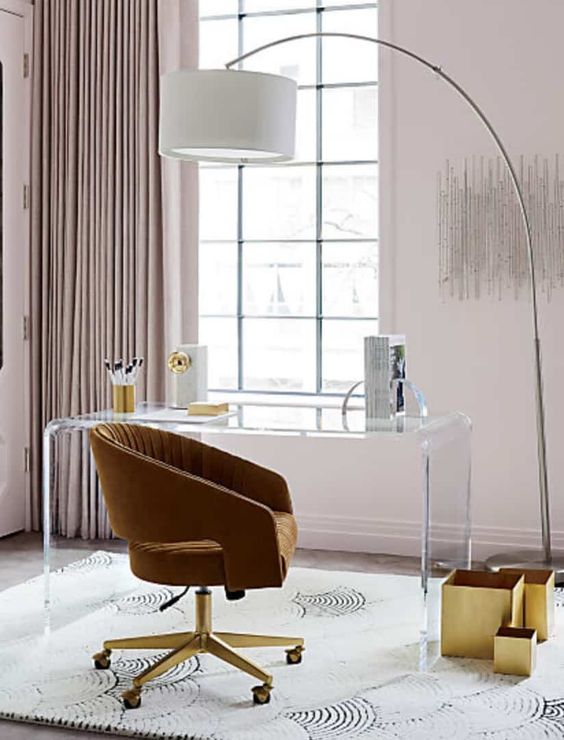 a gorgeous home office with an acrylic desk, a mustard chair, gold boxes, blush walls and matching curtains plus a floor lamp