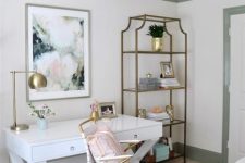 a lovely home office with a white desk, a white and gold chair, an open shelving unit, a gold chandelier and a floral artwork