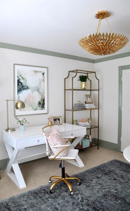 a lovely home office with a white desk, a white and gold chair, an open shelving unit, a gold chandelier and a floral artwork