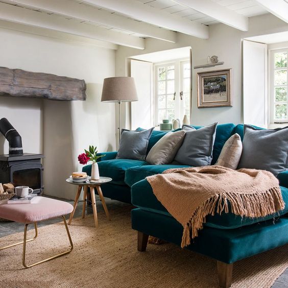 a lovely living room with a hearth with a wooden beam, a turquoise sofa, neutral textiles, a pink ottoman and a vintage lamp
