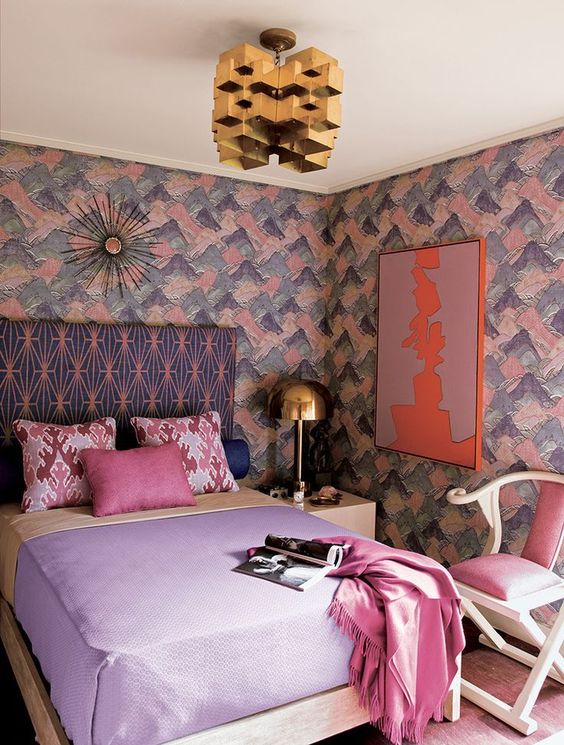 A lovely maximalist bedroom with brushstroke walls, a printed bed, pastel bedding, a pink chair, a gold sculptural chandelier