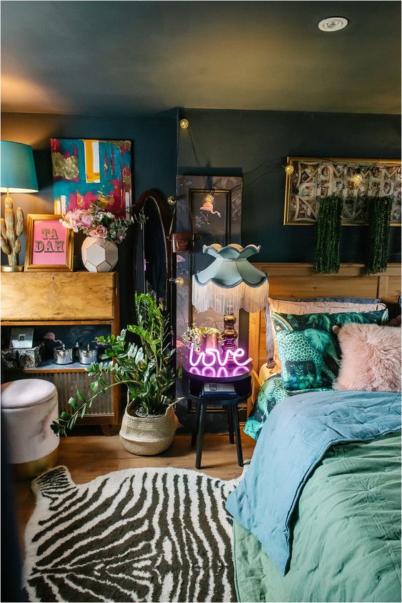 A lovely maximalist bedroom with dark walls and a ceiling, with mid century modern furniture, pastel bedding, a potted plant, a neon light and colorful artworks