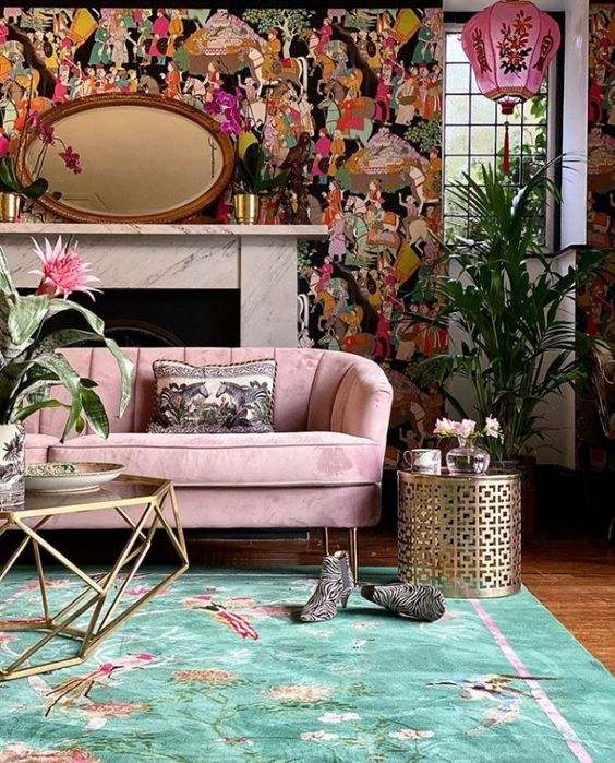 a lovely maximalist living room with a bold printed accent wall, a pink sofa, a green rug, metallic pieces and potted plants