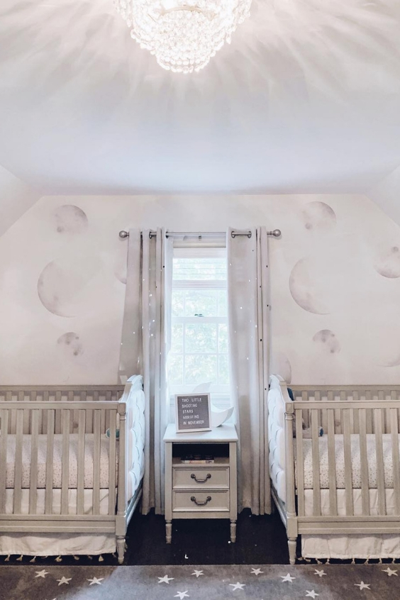 a lovely neutral nursery with lunar stenciled walls, grey cribs, a layered rugs and grey glitter curtains is very chic