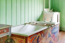 a maximalist bathroom with green planked walls, a colorful bathtub, a painted sink stand, pretty artworks and chic
