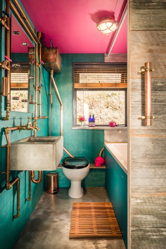 a maximalist bathroom with green walls and a hot pink ceiling, a concrete floor and a sink, copper exposed pipes and a skull