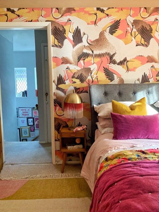 A maximalist bedroom with a bird printed wall, a grey bed, bright bedding and a rug, mid century modern nightstands
