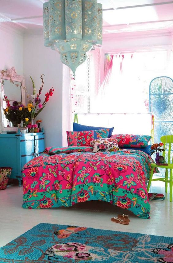 A maximalist bedroom with pink walls, a bed, a blue chest of drawers, a green chair, colorful bedding and bold blooms plus a lovely pendant lamp