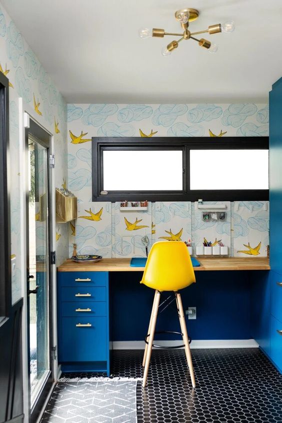 a maximalist home office with a penny tile floor, a bright blue desk, pretty printed wallpaper, a yellow stool and a gilded chandelier
