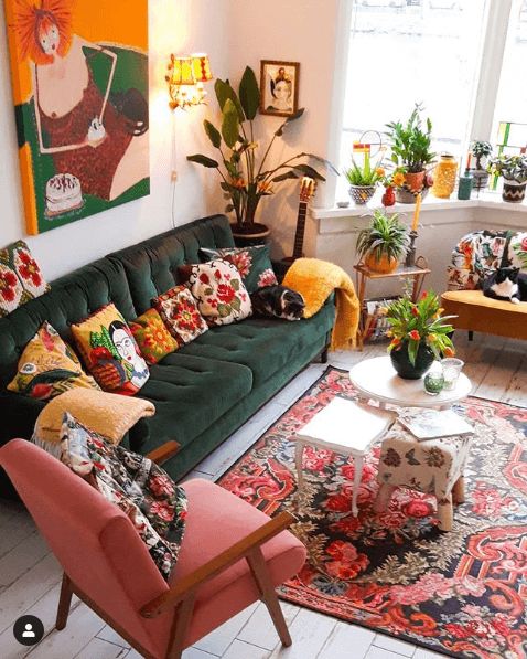 a maximalist living room with a green sofa and mustard and pink chairs, a bold rug and colorful pillows, potted plants and candleholders