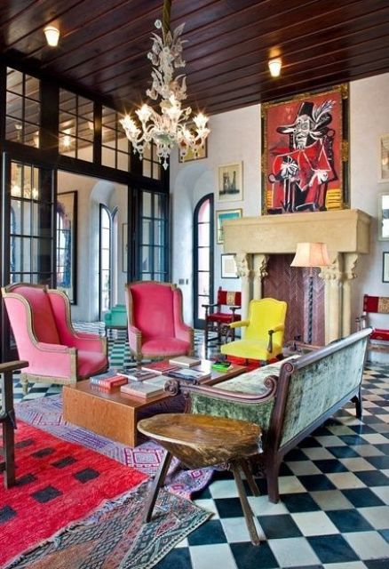 A maximalist living room with a non working fireplace and a checked floor, pink chairs, a green sofa and colorful rugs plus bold artworks