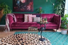 a maximalist living room with green walls, layered rugs, a fuchsia sofa, a bold artwork and potted greenery is chic