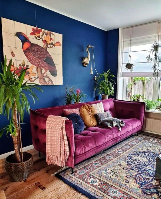 a maximalist living room with navy walls, a fuchsia sofa, a bold printed rug, potted plants and a lovely artwork is chic