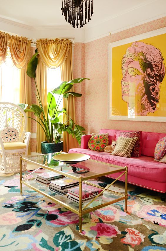 a maximalist living room with pink wallpaper walls, a hot pink sofa, a peacock chair, a glass coffee table, a colorful floral rug and yellow curtains