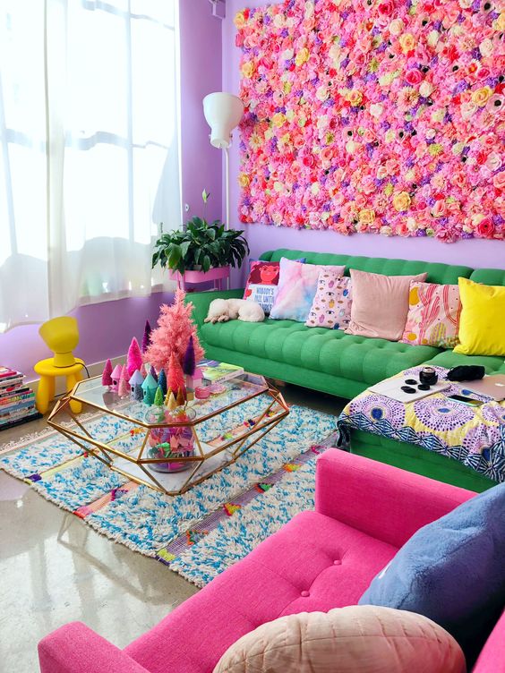 a maximalist living room with purple walls, a bright green and pink sofa, colorful pillows, a glass coffee table and a bright flower wall