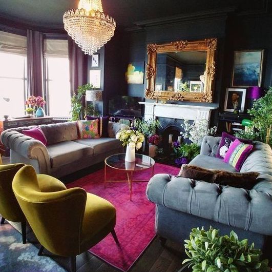 a moody maximalist living room with black walls, a bow window with purple curtains, a fireplace, grey sofas and a yellow chair