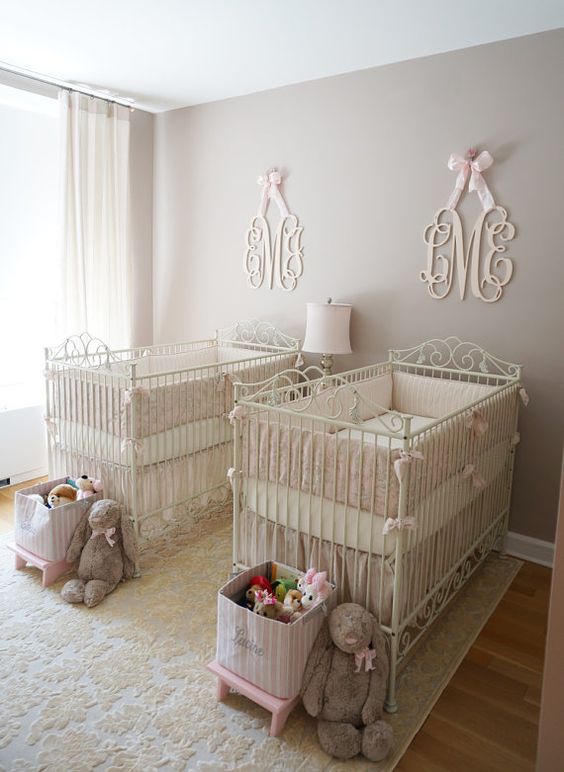 a neutral and pastel nursery with grey walls, white forged cribs, baskets for toys and refined monograms over each crib