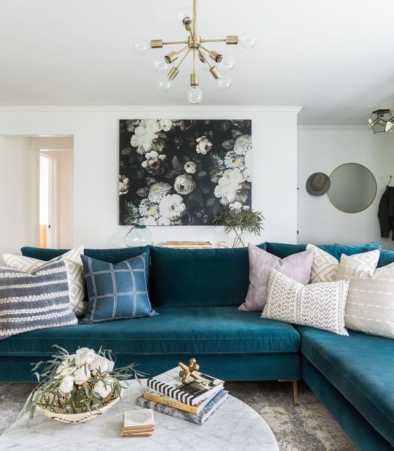 a pretty living room with a turquoise sectional, a dark floral artwork, a marble coffee table and a gold chandelier