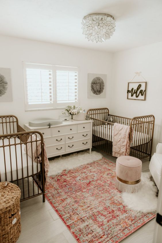 a pretty twin nursery with a white dresser and changing table, metal cribs, a Persian rug, a basket for storage and a pink ottoman