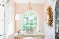 a refined and fancy home office with blush walls, a chic desk, a clear and upholstered stool and a large artwork