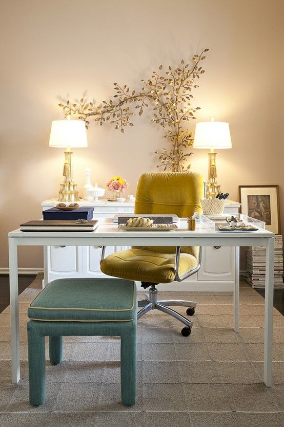 a refined and fancy home office with tan walls, a chic sleek desk, a mustard chair and a green stool, a gold leaf branch and matching lamps