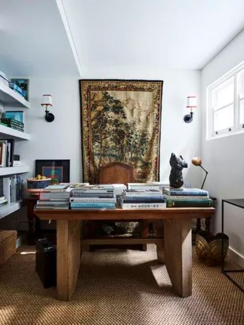 a refined maximalist home office with white walls, floating shelves, a rough wooden desk, a jute rug, baskets and tables and a tapestry
