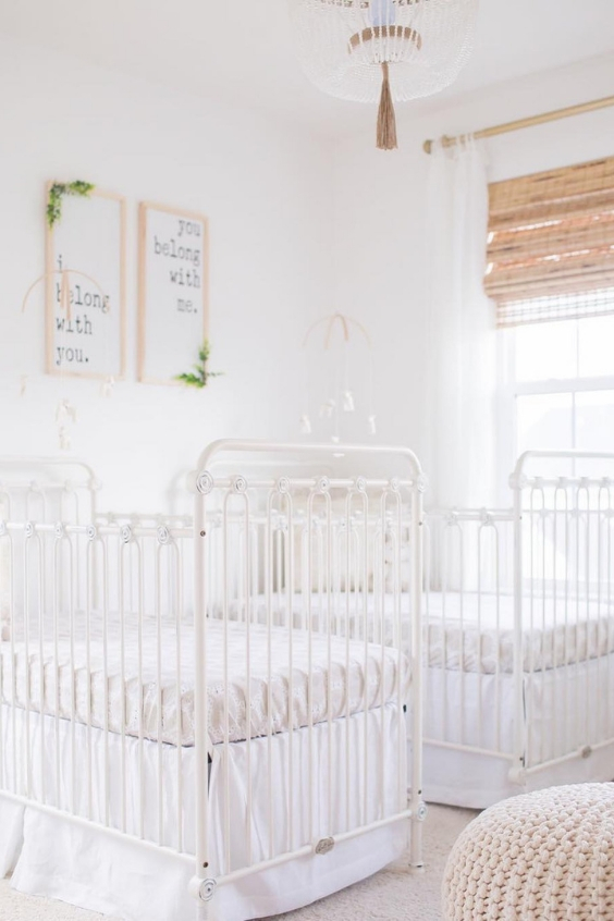 a serene white nursery with vintage white cribs, a knit ottoman, shades, artworks and a pretty chandelier plus matching mobiles