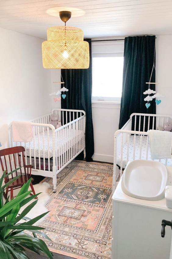 A small and cute twin nursery with white cribs, navy black out curtains, a printed rug, potted plants and a lovely retro lamp