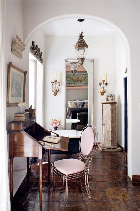 a sophisticated home office with a vintage bureau desk, a sheer blush chair, vintage lamps and candleholders on the walls
