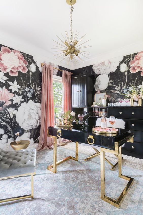a sophisticated home office with moody floral wallpaper, a black and gold desk, chic tufted chairs, black storage units and a sunburst chandelier